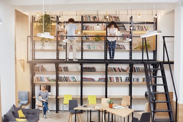 Foto gratuita phto de la gran biblioteca moderna de la universidad. chica rubia sentada en chear mirando en la ventana con expresión de la cara de ensueño. dos jóvenes de pie cerca de estanterías, leyendo libros.