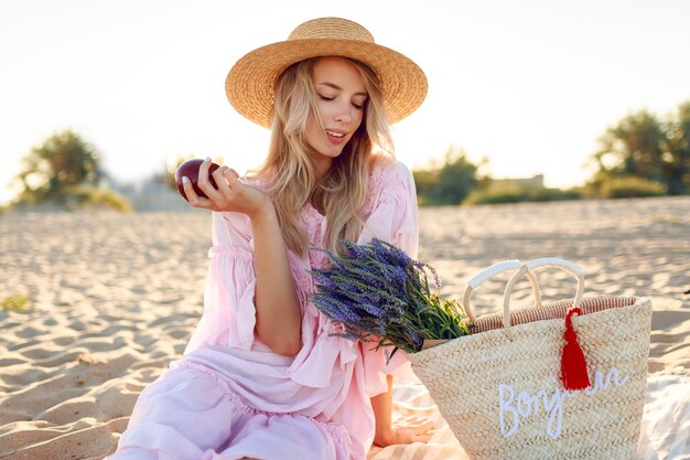 Picnic en el campo cerca del océano. Agraciada mujer joven con cabello rubio ondulado en elegante vestido rosa disfrutando de las vacaciones y comiendo frutas.
