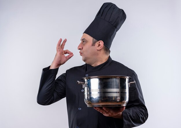 De pie en vista de perfil, cocinero masculino de mediana edad en uniforme de chef sosteniendo una cacerola mostrando un gesto delicioso