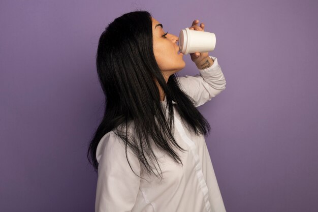 De pie en la vista de perfil hermosa joven vestida con camiseta blanca bebe café aislado en púrpura