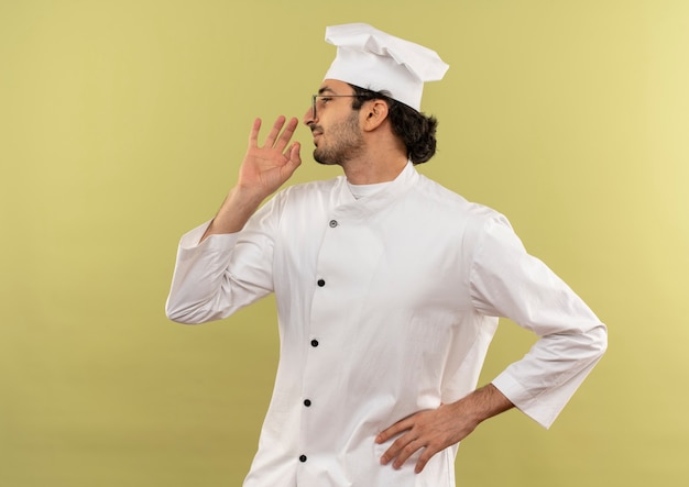 De pie en la vista de perfil, joven cocinero complacido con uniforme de chef y gafas mostrando un gesto delicioso y poniendo la mano en la cadera aislada en la pared verde