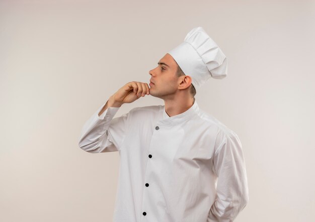 Foto gratuita de pie en la vista de perfil joven cocinero con uniforme de chef poniendo la mano en la barbilla con espacio de copia