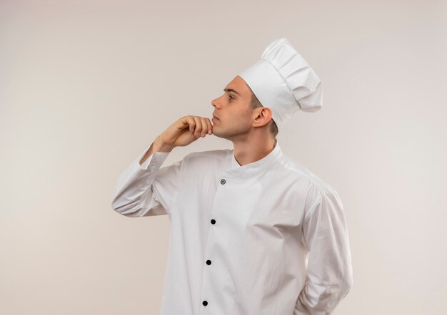De pie en la vista de perfil joven cocinero con uniforme de chef poniendo la mano en la barbilla con espacio de copia