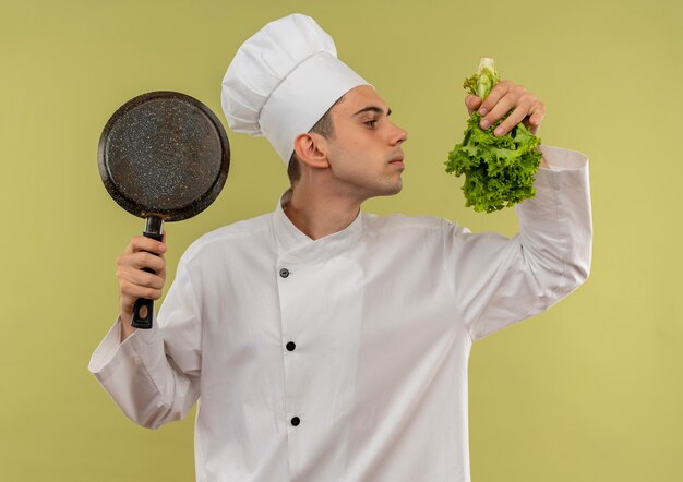 De pie en la vista de perfil joven cocinero vistiendo uniforme de chef sosteniendo una sartén oliendo ensalada en su mano