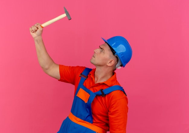 De pie en la vista de perfil joven constructor de sexo masculino vistiendo uniforme y casco de seguridad levantando y mirando el martillo