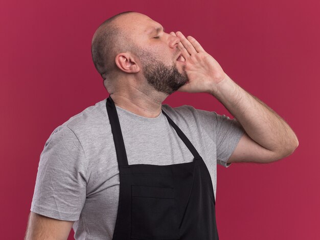 De pie en la vista de perfil con los ojos cerrados, barbero masculino de mediana edad eslavo en uniforme llamando a alguien aislado en la pared rosa