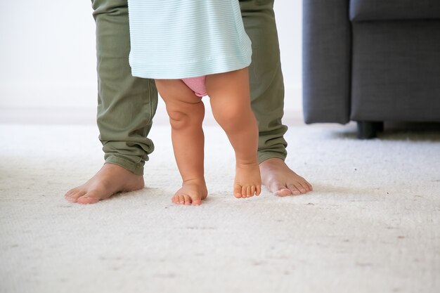 Piernas descalzas del bebé tratando de caminar delante de mamá. Niño dando sus primeros pasos con el apoyo de las mamás. Toma recortada. Concepto de paternidad e infancia