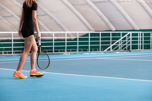 Piernas de joven en una cancha de tenis cerrada con pelota y raqueta