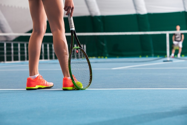 Piernas de joven en una cancha de tenis cerrada con pelota y raqueta