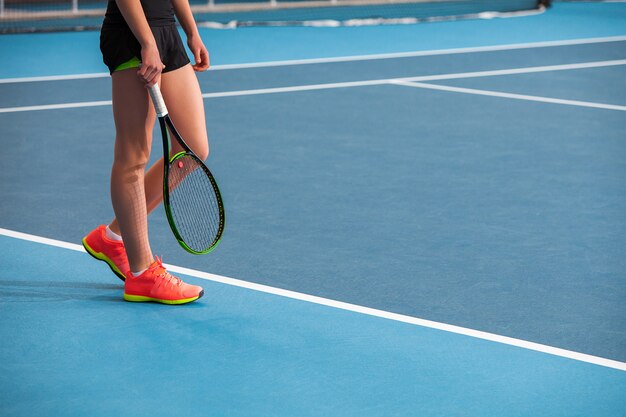 Piernas de joven en una cancha de tenis cerrada con pelota y raqueta