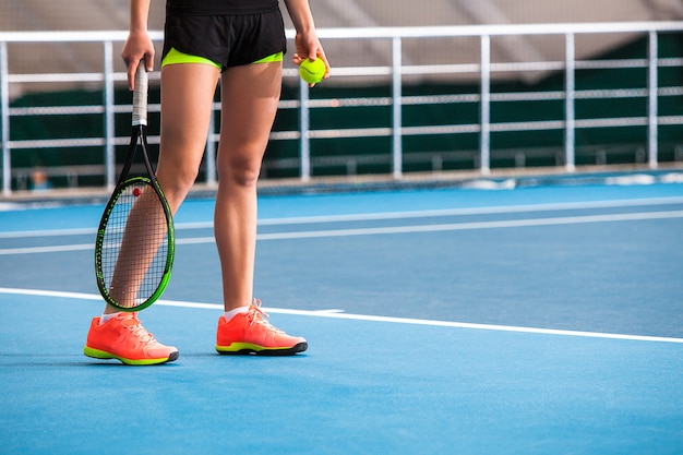 Piernas de niña en una cancha de tenis cerrada con pelota y raqueta