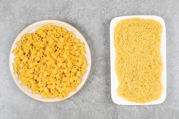 Pila de fideos sin cocer y pasta en un plato blanco sobre una mesa gris.