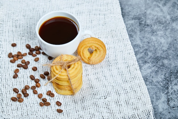Pila de galletas con granos de café y una taza de café sobre un mantel blanco.