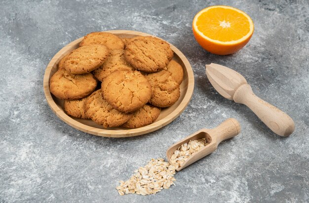 Pila de galletas sobre tabla de madera. Naranja medio cortada con avena sobre mesa gris.