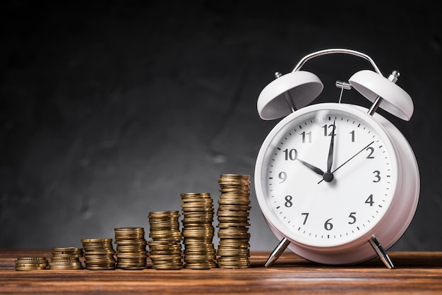 Pila de monedas cada vez mayores con el reloj de alarma blanco en el escritorio de madera contra fondo negro