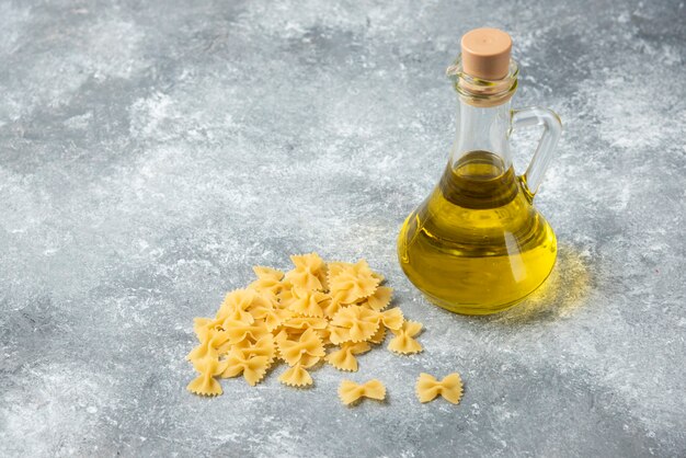 Pila de pasta farfalle cruda con una botella de aceite de oliva sobre fondo de mármol.