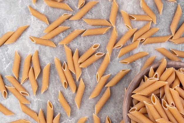 Pila de pasta penne marrón colocada en un recipiente de madera sobre una superficie de mármol.