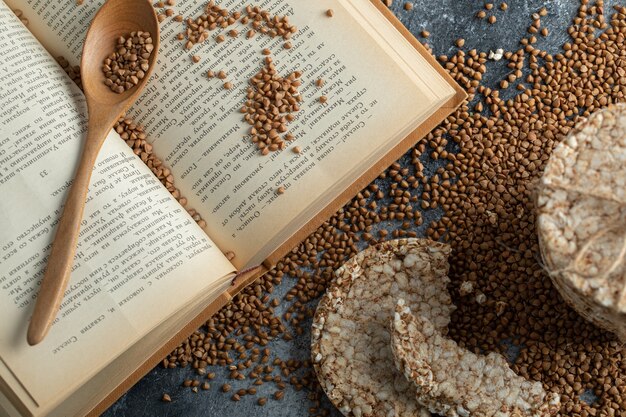 Pila de tortas de arroz, libro y trigo sarraceno crudo sobre superficie de mármol