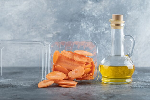 Pila de zanahorias en rodajas en un recipiente de plástico y una botella de aceite sobre fondo gris.