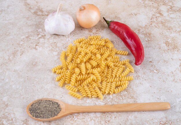 Foto gratuita pimienta negra en una cuchara de madera y un montón de pasta cruda cerca de un pequeño paquete de verduras en la superficie de mármol.