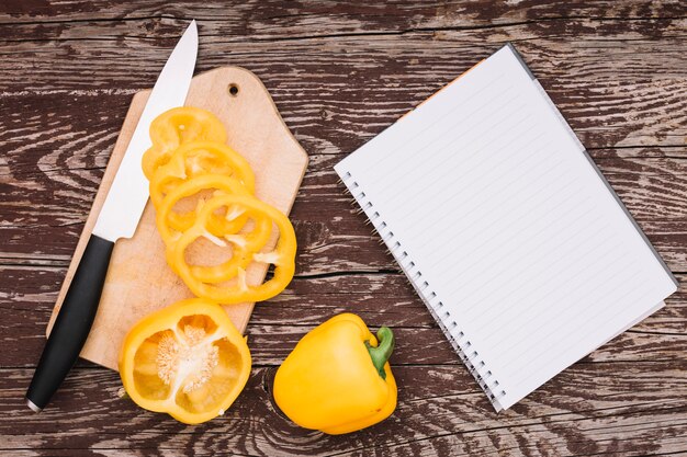 Pimiento amarillo entero y cortado en tabla de cortar con cuchillo y bloc de notas en espiral en el escritorio de madera