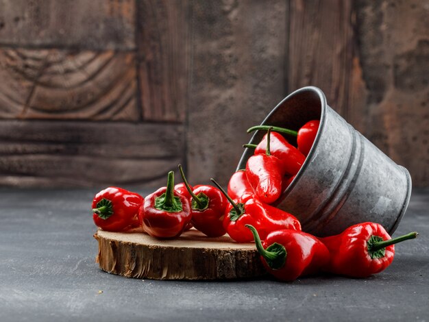 Pimientos rojos dispersos de un mini cubo con vista lateral de pieza de madera en pared de azulejos grises y piedra