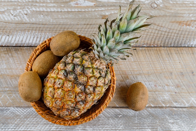 Piña en una cesta de mimbre con kiwis en una superficie de madera