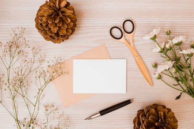 Piña; cortar con tijeras; flores de aster y aliento de bebé; Pluma y tarjeta en blanco en el escritorio de madera