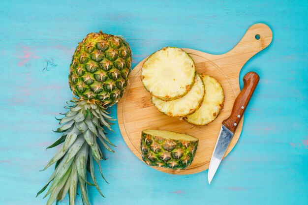 Piña entera y en rodajas con un cuchillo de frutas en una tabla de cortar en azul cian, endecha plana.