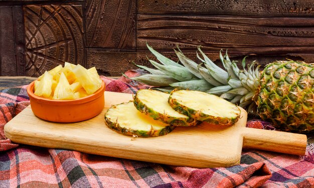 Piña entera y en rodajas en una tabla de cortar y un tazón de arcilla en un azulejo de piedra y tela de picnic. vista lateral.