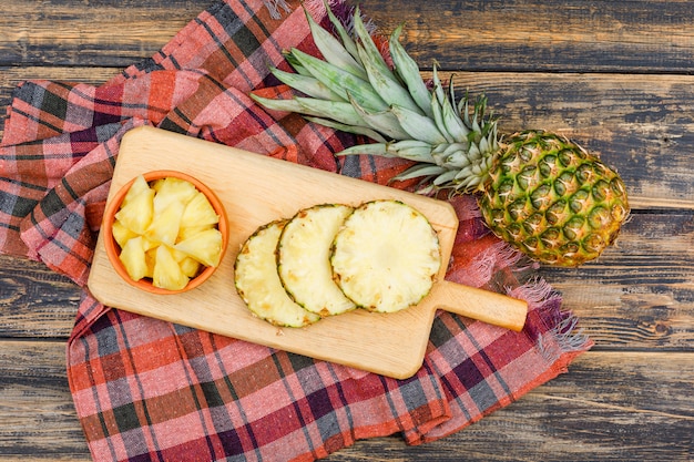 Piña entera y en rodajas en una tabla de cortar y un tazón de arcilla sobre una superficie de madera grunge y tela de picnic. aplanada