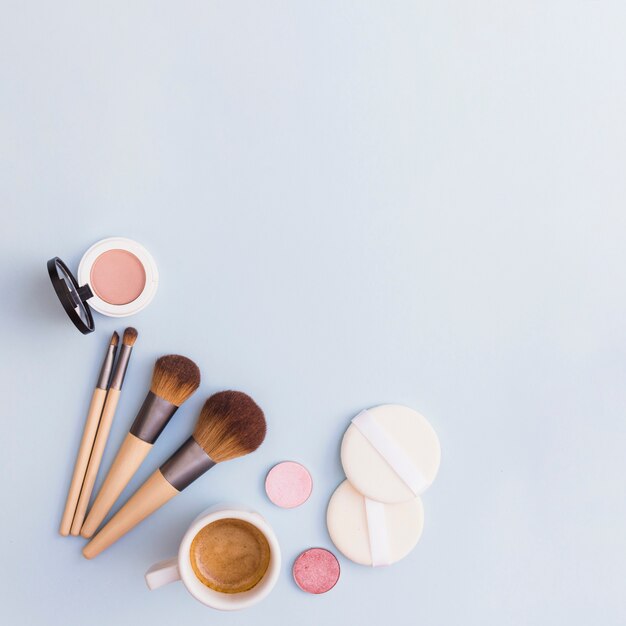 Pinceles de maquillaje; sombra; Colorete y esponja con taza de café sobre fondo azul