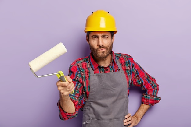 El pintor serio sostiene el rodillo de pintura, hace la redecoración en casa, pinta las paredes, usa casco protector y delantal, posa en el interior, ocupado con la reparación y renovación, aislado en la pared púrpura.