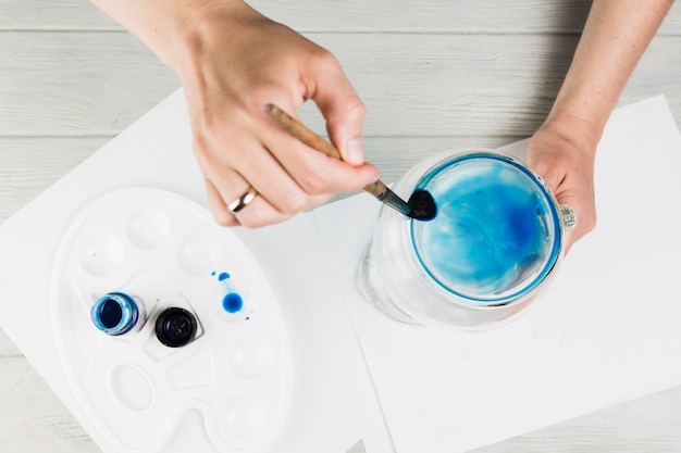 Foto gratuita pintura femenina de la mano en el tarro de cristal con el cepillo de pintura sobre el escritorio de madera