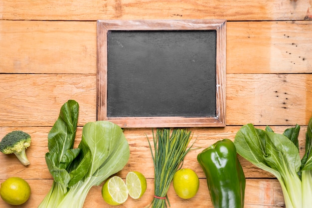 Pizarra en blanco con hilera de bokchoy; brócoli; limón; pimiento; cebolletas en el escritorio de madera