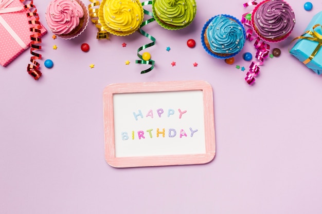 Pizarra de feliz cumpleaños con gemas de colores; serpentinas y muffins sobre fondo rosa