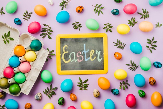 Pizarra con el título de Pascua entre el brillante conjunto de huevos de colores y hojas cerca del contenedor