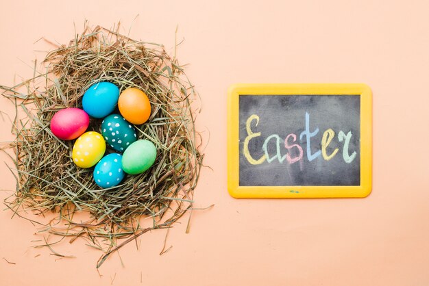 Pizarra con el título de Pascua cerca de conjunto de huevos de colores brillantes en el nido