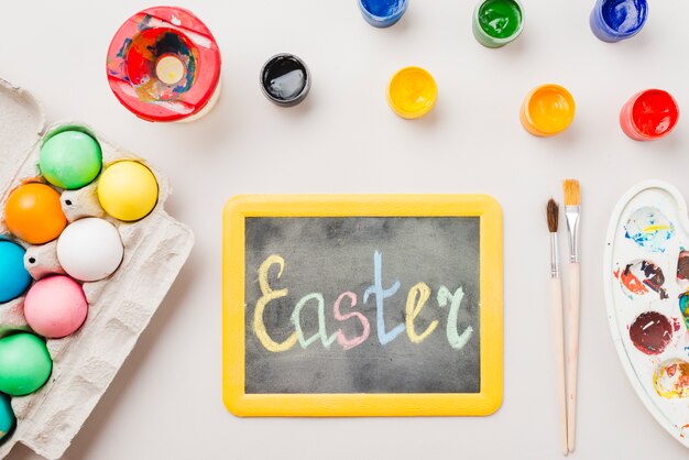 Pizarra con título de Pascua cerca de huevos de colores en contenedor, pinceles, acuarelas y paleta