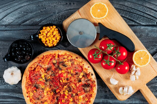 Pizza con ajo, tomate, limón, aceitunas, maíz y un cortador de pizza vista superior sobre un fondo de madera oscura