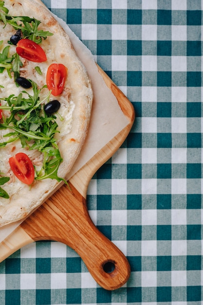 Foto gratuita pizza napolitana con atún, queso, rúcula, albahaca, tomates, aceitunas, espolvoreada con queso sobre una mesa de madera sobre un mantel en una celda con un lugar para el texto.