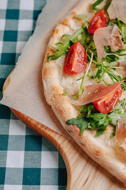 Pizza napolitana con jamón, queso, rúcula, albahaca, tomates espolvoreados con queso sobre una tabla de madera sobre un mantel en una celda con un lugar para el texto