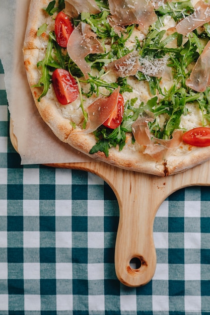 Foto gratuita pizza napolitana con salami, rúcula, tomates espolvoreados con queso sobre una tabla de madera sobre un mantel en una celda con un lugar para el texto