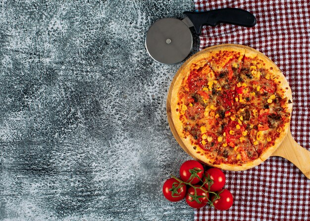 Pizza en una tabla de cortar con tomates, cortador de pizza vista superior sobre un fondo de tela de estuco gris y picnic