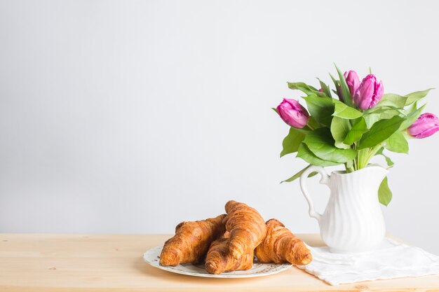 Placa de los croissants cocidos cerca del florero en el escritorio de madera aislado en el fondo blanco