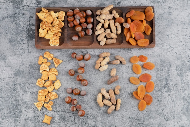 Placa de madera llena de nueces, galletas y albaricoques secos sobre superficie de mármol.