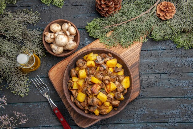 Placa de vista superior a bordo plato de patatas champiñones sobre tabla de madera junto al tenedor debajo del tazón de aceite de champiñones en botella y ramas de árboles con conos