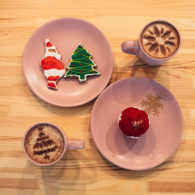 Placas de color rosa con dulces de Navidad de pie entre las tazas con café en la mesa de madera