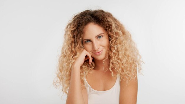 Plan de cabeza y hombros de mujer rubia con pelo rizado grande en estudio en blanco mirando a la cámara