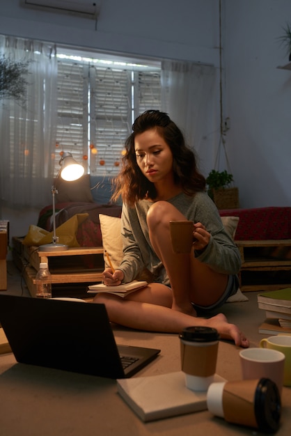 Plano completo de una estudiante asiática que se prepara para los exámenes por la noche sentada en el piso de su habitación con muchas tazas de café vacías